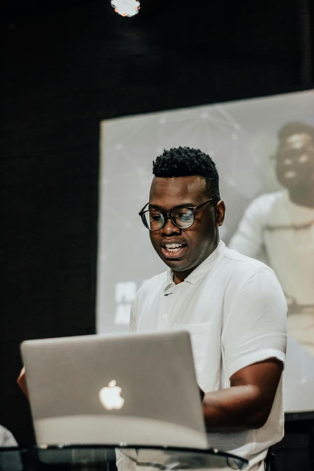 A man in a white shirt presenting in front of a laptop.